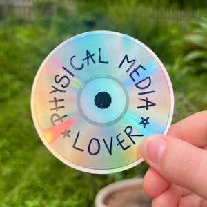 May include: A holographic CD sticker with the words "PHYSICAL MEDIA LOVER" in black, arranged around the edge. The sticker has a rainbow sheen and a black center, held against a blurred green background.