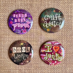 Op de afbeelding: Vier ronde knutselknoppen met tekst. Eén knop zegt "scrapbook princess" in paars. Een andere zegt "craft addict" in groen. Een derde zegt "book worm" in bruin. De laatste zegt "I love junk" in roze.