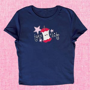 May include: A navy blue t-shirt with a pink and red embroidered apple with the text "take a bite" on it.