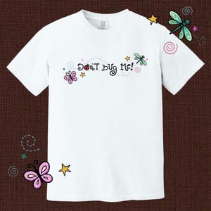 Op de afbeelding: Een wit t-shirt met korte mouwen en de tekst "Don't bug me!" in zwarte letters. Het ontwerp bevat kleurrijke illustraties van een roze vlinder, een rood lieveheersbeestje, een groene libel, gele sterren en kleine roze en blauwe krullen.
