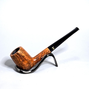 Puede incluir: Una pipa de tabaco con un cuenco marrón texturizado y una boquilla negra. La pipa descansa sobre un soporte plateado. La palabra "Stanwell" está impresa en el lateral de la pipa.