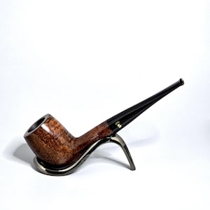 Puede incluir: Una pipa de tabaco marrón y negra con un cuenco curvado y un tallo largo y delgado. La pipa descansa sobre un soporte plateado sobre un fondo blanco. La pipa tiene un aspecto de veta de madera texturizada.