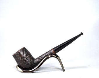 Op de afbeelding: Een bruine tabakspijp met een zwart mondstuk, rustend op een zilverkleurige pijpenstandaard. De pijp heeft een getextureerde ketel en een gladde, taps toelopende steel. De standaard heeft een gebogen ontwerp.