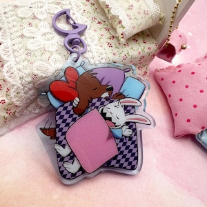 May include: A clear acrylic keychain featuring a cartoon dog and rabbit sleeping in a bed with pillows and a blanket. The keychain has a purple clasp and is surrounded by miniature bed and pillows.