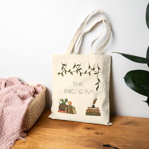 May include: A cream-colored canvas tote bag featuring a printed design of green ivy vines, a small green dragon perched on a stack of books, and a quill pen in an inkwell on another stack of books with the text THE INK & IVY printed in black.
