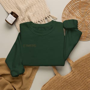 May include: A dark forest green long-sleeve crewneck sweatshirt featuring the word ESMERE embroidered in a thin, gold-coloured font on the upper left chest area. The garment has a ribbed collar, cuffs, and hem, with a simple, clean design.