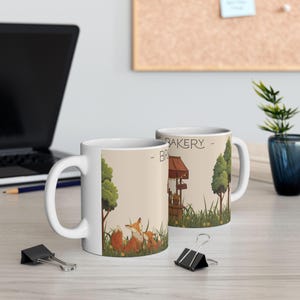 May include: A white ceramic mug featuring a printed illustration of two foxes sitting in a grassy field near a small bakery stand. The text on the mug reads THE BAKERY. The design includes green trees, yellow flowers, and a brown wooden stall structure.
