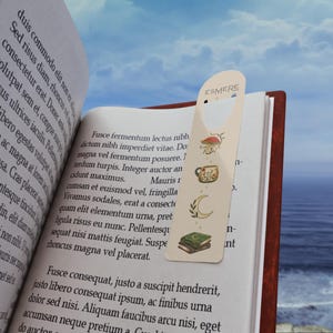 May include: A cream-coloured bookmark with a die-cut top featuring the text ESMERE. The bookmark displays vertical illustrations of a mushroom, a floral mug, a crescent moon with leaves, and a stack of two books with a moon symbol on the cover.
