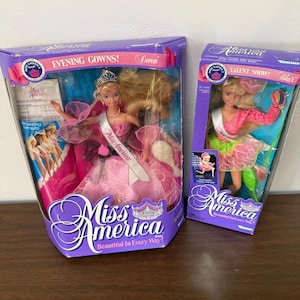 Può includere: Due bambole Miss America nella confezione originale. La scatola a sinistra mostra Devon in un abito da sera rosa con tiara e fascia. La scatola a destra mostra Blair in un completo da spettacolo rosa e verde. Entrambe le scatole hanno il logo.