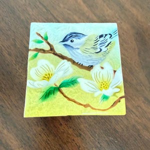 Puede incluir: Un azulejo decorativo cuadrado con una escena pintada a mano de un pájaro pequeño con plumaje azul y amarillo posado en una rama marrón. Dos flores de cornejo blancas con centros amarillos y hojas verdes se sitúan bajo el pájaro sobre fondo amarillo.