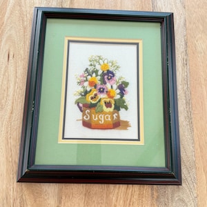 Pode incluir: Bordado floral emoldurado com um buquê de amores-perfeitos e outras flores em uma açucareira. A palavra "Sugar" é bordada na açucareira. A moldura é de madeira com uma passe-partout verde.