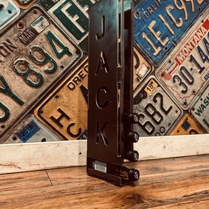 May include: Black metal bottle holder with the word "JACK" cut out. The holder is designed to hold six bottles of liquor. The holder is sitting on a wooden surface.