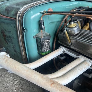Może przedstawiać: Komora silnika zabytkowego turkusowego samochodu z przymocowaną butelką whisky Jack Daniel's. Widoczne miedziane rury i kolektory wydechowe. Nadwozie samochodu ma wygląd postarzałego i zużytego.