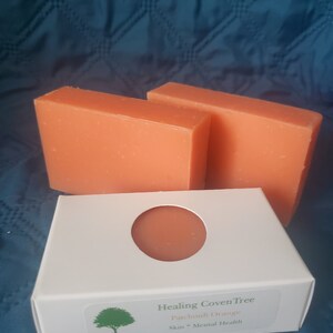 May include: Two orange bars of soap in a white box with a cutout for the soap. The box has a green tree logo and the text "Healing Coven Tree Patchouli Orange Skin - Mental Health".