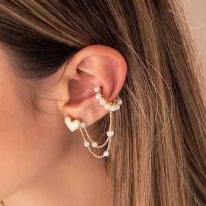 May include: Gold ear cuff with a delicate chain and pearl accents. The ear cuff is shaped like a crescent moon and has a small heart charm attached to the chain.