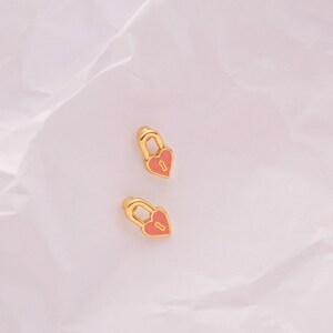 May include: A pair of gold heart-shaped lock earrings with pink enamel. The earrings have a small keyhole detail.