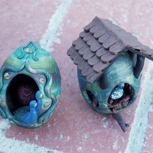 Puede incluir: Dos pajareras artesanales. Una tiene forma de huevo con un pavo real y un caracol en su interior. La otra es de diseño tradicional con tejado marrón y un huevo azul. Ambas pintadas en tonos azules, verdes y marrones.