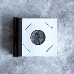 Puede incluir: Un centavo de Lincoln de 1987 en un soporte blanco para monedas con lomo negro. La moneda presenta un perfil de Abraham Lincoln y la palabra "LIBERTY". El soporte está asegurado con grapas metálicas.