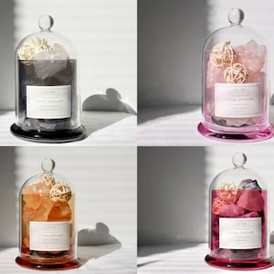 May include: Four glass cloche jars with clear lids, each containing different colored crystals and decorative elements. The jars have black and pink bases and feature a label that reads "CIUNA Crystal Popurrí".