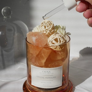 May include: A glass jar filled with pink Himalayan salt crystals, white wicker balls, and white flowers. The jar has a label that reads "CIUNA AMBIENCE CREATOR CRYSTAL POTPOURRI NATURAL CRYSTAL". A hand is holding a dropper bottle and adding liquid to the jar.