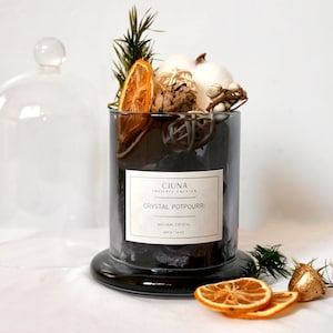 May include: A black glass jar filled with a crystal potpourri, with dried orange slices, pine sprigs, white cotton balls, and white berries. The jar has a black label that reads "CIUNA" and "CRYSTAL POTPOURRI".