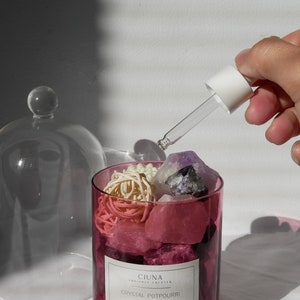 May include: A clear glass jar with a lid filled with pink and purple crystals and white dried flowers. A glass dropper bottle with a white cap is being used to add essential oil to the jar. The jar has a label that reads "CIUNA AROMATIC CREATOR CRYSTAL POTPOURRI".