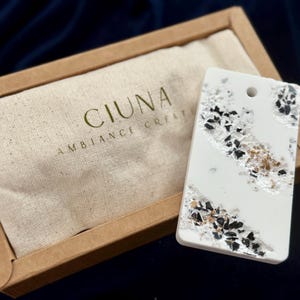 May include: A white rectangular decorative item with black, silver, and gold accents, presented in a brown box with a beige fabric lining. The fabric is printed with the text "CIUNA AMBIANCE CREAT".