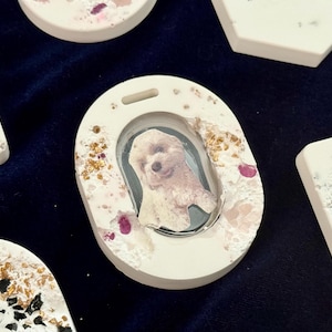 May include: Oval-shaped white pet memorial with a photo of a dog, surrounded by gold flakes and pink accents. Other white, decorative pet memorials are also visible, each with unique designs and shapes.
