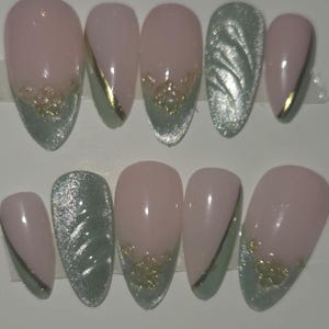 May include: Set of ten oval-shaped false nails. The nails feature a light pink base with accents of silver glitter, gold glitter, and a silver-green color. Some nails have a gold stripe or glitter design.