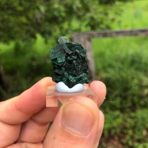 Op de afbeelding: Een donkergroen malachiet mineraal specimen. Het mineraal is gemonteerd op een heldere acryl basis met een witte steun. De achtergrond is wazig en toont groen gebladerte en een houten hek.