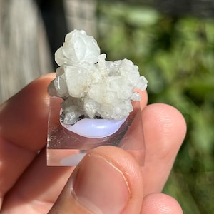 Puede incluir: Un grupo de cristales translúcidos y de color claro se asienta sobre una base cuadrada transparente con un acento ovalado azul claro. Los cristales tienen un aspecto rugoso y texturizado. El objeto se sostiene en la mano de una persona.