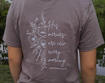 May include: A taupe-colored t-shirt with a floral design and the text "His mercies are new every morning" in white script. The shirt also includes the Bible verse Lamentations 3:22-23.
