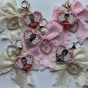 May include: Five heart-shaped acrylic keychains featuring portraits of soccer players Cristiano Ronaldo, Bellingham, Haaland, and others. Each keychain includes a pink and white checkered background, a red ribbon border, a pearl-accented heart charm, and a fabric bow.