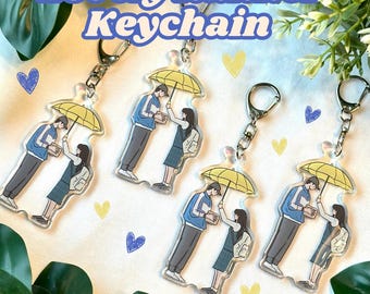 May include: Four clear acrylic keychains featuring a cartoon couple holding an umbrella. The couple is dressed in blue and gray clothing. The keychains are yellow, blue, and gray.