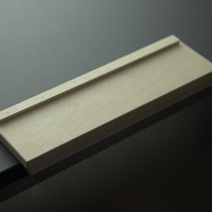 Könnte beinhalten: Ein rechteckiges Holztablett mit erhöhter Kante und kleinen Löchern. Das Tablett besteht aus hellem Holz und liegt auf einer dunklen Oberfläche. Das Design ist schlicht und minimalistisch.