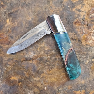 Puede incluir: Una navaja de bolsillo plegable con una hoja de acero estampada y un mango con tonos de verde azulado, marrón y dorado. El cuchillo tiene una virola plateada y un acento azul. La hoja está parcialmente abierta, revelando su intrincado diseño.