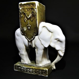 Op de afbeelding: Witte keramische olifantfiguur met een gouden en bruin gepatroneerd zadel. De olifant staat op een voet met een gouden en bruin gepatroneerde rand.