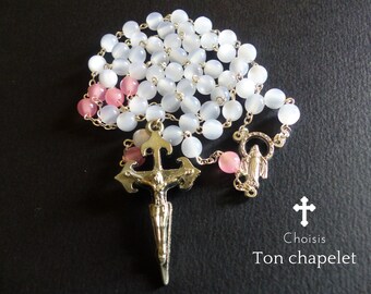 May include: White and pink beaded rosary with a silver cross and a silver charm of the Virgin Mary. The rosary is on a black background. The text "Choisis Ton chapelet" is visible below the rosary.