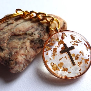 Könnte beinhalten: Goldener Schlüsselanhänger mit einem runden Harz-Anhänger mit einem goldenen Kreuz und goldenen Flocken. Der Anhänger ist an einer goldenen Kette und einem goldenen Schlüsselring befestigt.