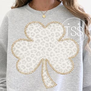 May include: A light grey sweatshirt with a large shamrock design. The shamrock has a white and grey leopard print and gold stitching. A casual top for St. Patrick's Day.