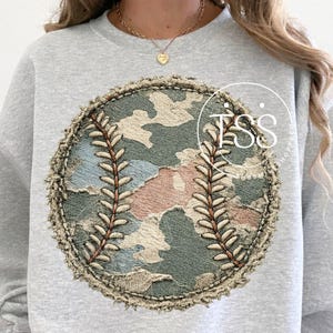 Puede incluir: Una sudadera gris claro con un diseño de béisbol. El gráfico de béisbol presenta un patrón de camuflaje en tonos verdes, rosas y azules. El béisbol está detallado con costuras marrones y un borde texturizado y desgastado.