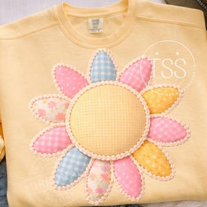 Peut inclure: Un sweat-shirt jaune clair orné d'une grande fleur appliquée. La fleur a un centre en vichy jaune et des pétales roses, bleus à carreaux, jaunes à pois et à motifs floraux. Le texte "TSS SHOPPE CO." est visible.