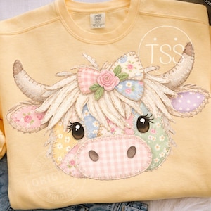 May include: A light yellow sweatshirt featuring a patchwork cow face design. The cow has a pink gingham nose, floral patches, and a pink bow with a rose. The sweatshirt is a Comfort Colors brand.
