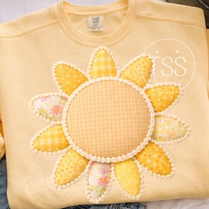 Peut inclure: Un sweat-shirt jaune clair orné d'un grand motif de tournesol. Le tournesol a un centre en vichy et des pétales avec divers motifs jaunes, dont du vichy et des imprimés floraux. Le motif est souligné d'une bordure blanche.
