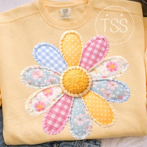 Peut inclure: Un sweat-shirt jaune clair orné d'une grande marguerite appliquée. Les pétales de la marguerite présentent différents motifs : vichy bleu, vichy rose, floral et pois jaunes. Le centre de la fleur est un cercle jaune texturé.