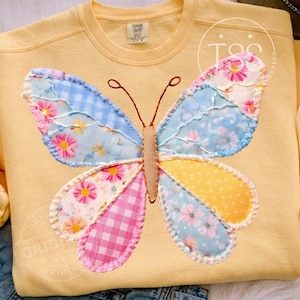 May include: A yellow sweatshirt featuring a large, appliqued butterfly. The butterfly's wings are composed of various patterned fabrics in blue, pink, and yellow, with floral and gingham designs. The sweatshirt has long sleeves and a crew neck.
