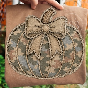 May include: A tan t-shirt featuring a large, embroidered pumpkin design. The pumpkin has a camouflage pattern in shades of green, brown, and gray, with a textured tan bow at the top. The entire pumpkin motif is outlined with a decorative stitch.