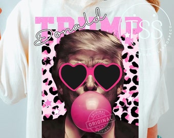 May include: Cream-colored t-shirt featuring a graphic of a man with heart-shaped sunglasses blowing a bubblegum bubble. The word "TRUMP" is printed in pink above the image, with "Donald" in cursive. A pink leopard print and the word "ORIGINAL" are also visible.
