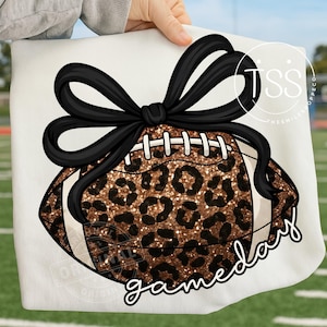 Puede incluir: Una sudadera blanca con un diseño de fútbol americano con estampado de leopardo y un lazo negro. Las palabras "gameday" están escritas en cursiva. El diseño es sostenido por una persona.
