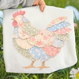 Könnte beinhalten: Ein weißes Stoffstück mit einem Patchwork-Hühnerdesign. Das Huhn besteht aus verschiedenen pastellfarbenen Stoffen mit Blumen- und Punktmustern. Es hat einen rosa Kamm und Schnabel, mit braunen Füßen. Der Artikel wird vor einem grasbewachsenen Hintergrund gehalten.